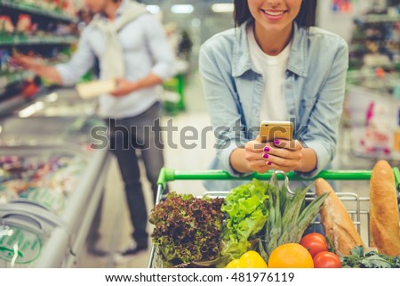 Couple in the supermarket. Cropped image of girl leaning on shopping cart, using a mobile phone and smiling, in the background her boyfriend is choosing food