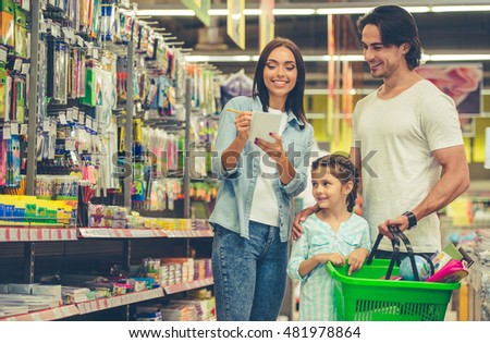 Beautiful young parents and their cute little daughter are smiling while choosing school stationery in the supermarket. Mom is making notes in the list
