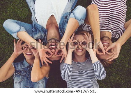 Top view of beautiful stylish friends making glasses of fingers, looking at camera and smiling while lying on grass