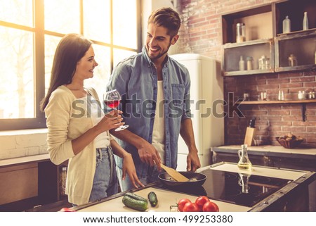 Beautiful young couple is talking and smiling while cooking in kitchen at home. Woman is drinking wine while her man is frying food