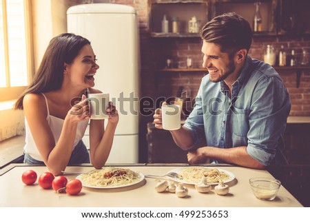 Beautiful young couple is talking, looking at each other and smiling while eating and drinking in kitchen at home