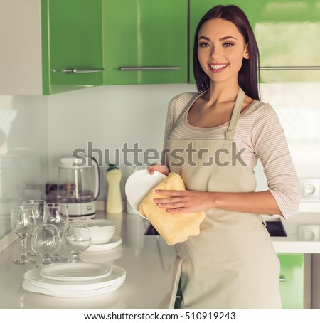 Beautiful young woman in apron is wiping the dishes, looking at camera and smiling while cleaning in kitchen