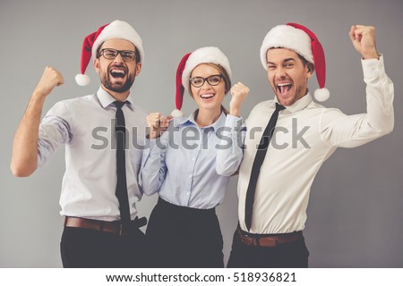 Happy business people in Santa hats are looking at camera and smiling while celebrating New Year, on gray background