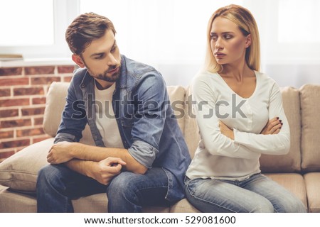 Beautiful couple is sitting back to back on the couch and looking offended at each other while having a quarrel