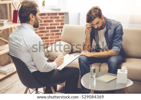 Handsome young man is sitting on couch and talking to the psychologist while doctor is making notes