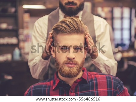 Handsome bearded man is looking at camera while getting haircut by hairdresser at the barbershop
