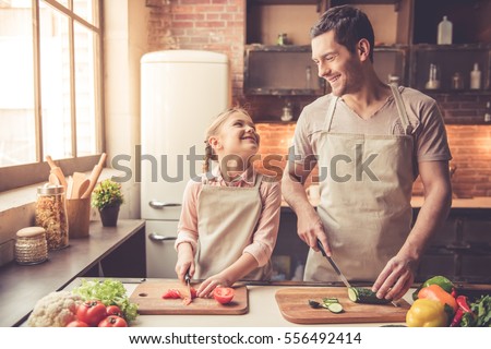Cute little girl and her handsome dad are cutting vegetables and smiling while cooking in kitchen at home