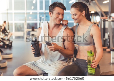 Attractive young sports people are holding bottle of water, talking and smiling while resting in gym