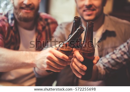 Cropped image of handsome friends clinking bottles of beer and smiling while resting at the pub