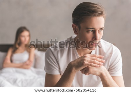 Young couple having a problem. Guy is sitting on bed and looking sadly away, his girlfriend in the background