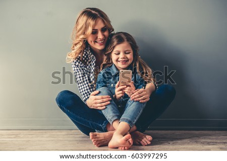 Cute little girl and her beautiful young mom are sitting together on the floor, using a smart phone and smiling, on gray background