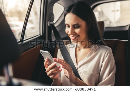 Beautiful business woman is using a smart phone and smiling while sitting on back seat in the car