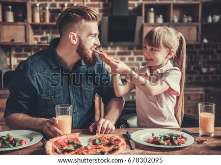 Cute little girl and her handsome bearded dad are smiling while drinking juice and eating pizza in kitchen