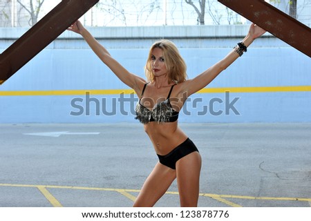 Tall blond woman posing very sexy in bikini in a parking lot garage exterior.