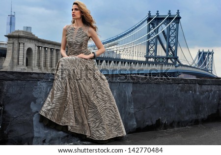 Fashion model posing sexy, wearing long evening dress on rooftop location with metal bridge construction on background