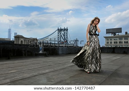 Fashion Model Posing Sexy, Wearing Long Evening Dress On Rooftop Location With Metal Bridge Construction On Background
