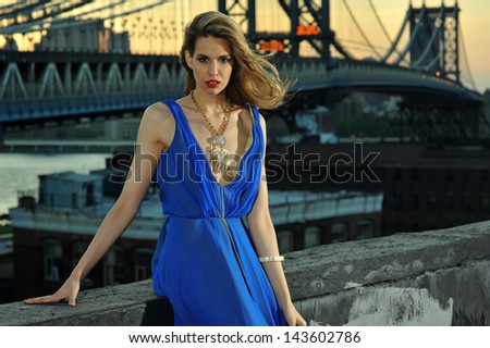 Fashion model posing sexy, wearing long blue evening dress on rooftop location with metal bridge construction on background