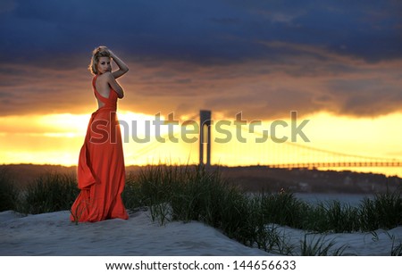 Young fashion model posing in red dress on sunset time with effective background of clouds and Verrazano bridge