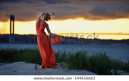 Young fashion model posing in red dress on sunset time with effective background of clouds and Verrazano bridge