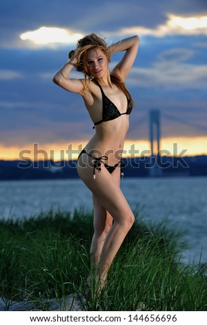 Young fit model posing in bikini on sunset time with effective background of clouds and Verrazano bridge