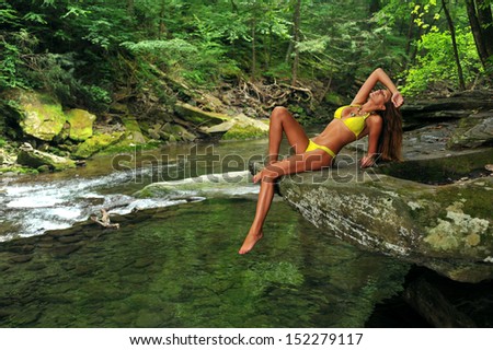 Sexy young woman posing in designer bikini at exotic location of mountain river with green water, rocks and forest in background