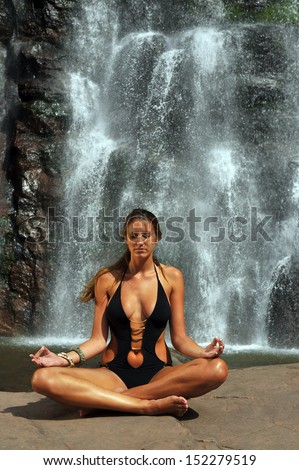 Beautifull girl wearing black one piece swimsuit meditating in lotus yoga pose in front of waterfall