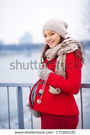Winter outdoor portrait of pregnant woman in fashionable clothes standing by the river