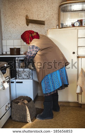 Very old woman is putting wood in her old wood-burning stove in her country style kitchen