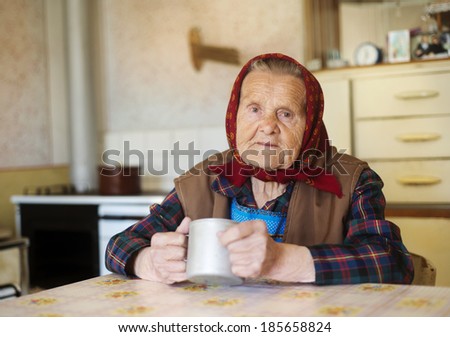 Very old woman in head scarf is drinking tea in her old country style kitchen