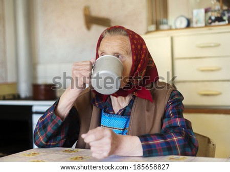Very old woman in head scarf is drinking tea in her old country style kitchen
