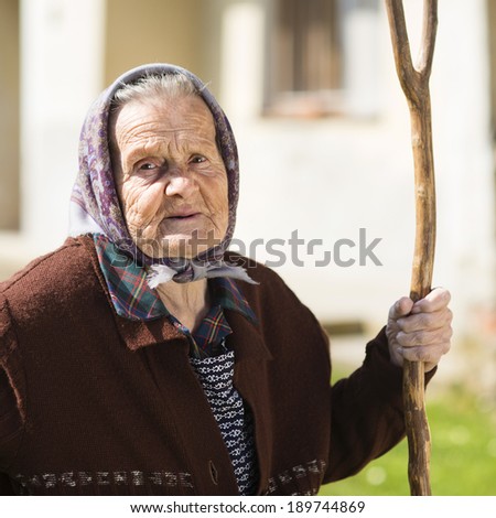 Very old woman in head scarf with rake in her backyard