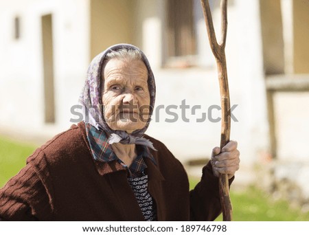 Very old woman in head scarf with rake in her backyard