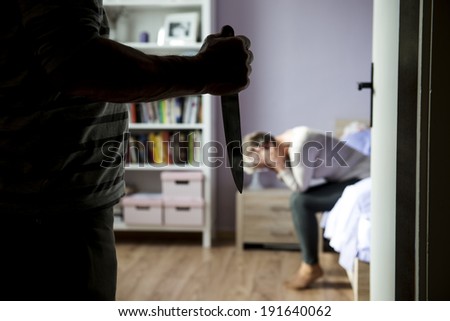 Mature woman siiting on the bed is scared of man with knife. Woman is victim of domestic violence and abuse.