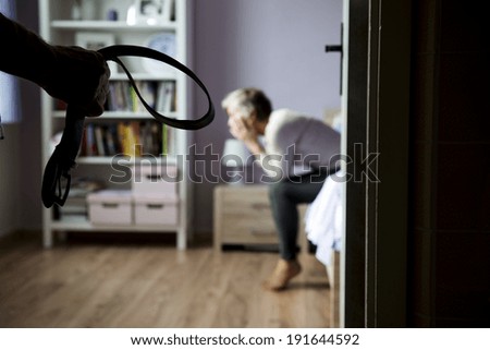 Mature woman siiting on the bed is scared of man with belt. Woman is victim of domestic violence and abuse.
