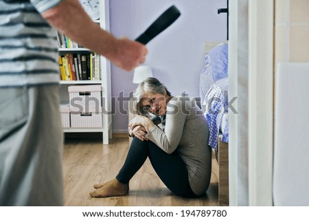 Mature woman siiting on the floor is scared of man with belt. Woman is victim of domestic violence and abuse.