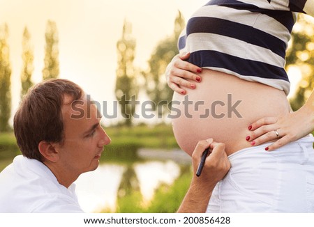 Man is writing a name on pregnant woman\'s belly in nature