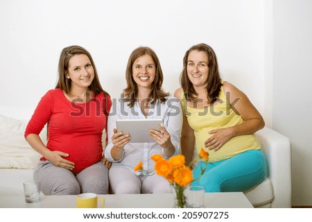 Three pregnant women sitting on sofa, chatting and using digital tablet