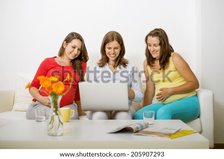 Three pregnant women sitting on sofa, chatting and using laptop