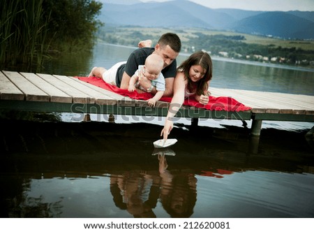 Mother and father with baby boy sitting on pier and playing with paper boat