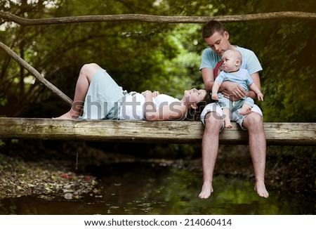 Happy family with little baby boy is sitting on the bridge over the river