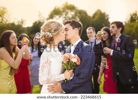 Young newlyweds kissing and enjoying romantic moment together at wedding reception outside, wedding guests in background blowing bubbles