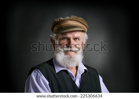Portrait of old bearded man in traditional cap, posing in studio on black background