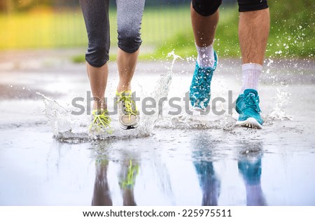 Young couple running on asphalt sports field in rainy weather. Details of legs and sports shoes splashing in puddles.