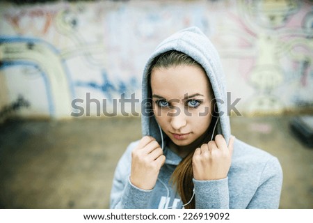 Serious teenage girl with hood on listening to music from headphones standing on background of graffiti wall