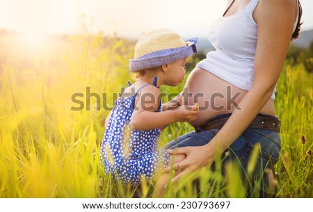 Cute little girl hugging and kissing her mother\'s pregnant belly in summer nature
