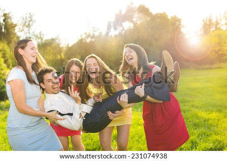 Funny portrait of bridesmaids holding groom on their hands in green sunny park