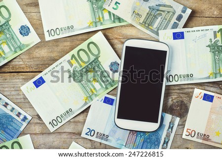 Smartphone and European currency banknotes scattered on the wooden table background. View from above.