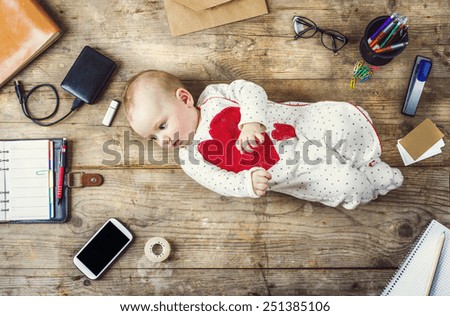 Mix of office supplies and gadgets on a wooden desk background alongside with cute little baby. View from above.