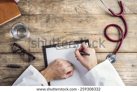 Hands of unrecognizable doctor writing on a blank sheet of paper. Wooden desk background.