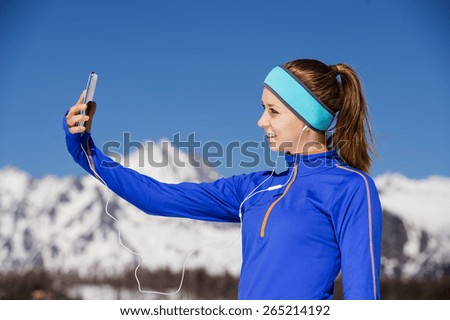 Young sportswoman jogging outside in sunny winter mountains taking selfie.